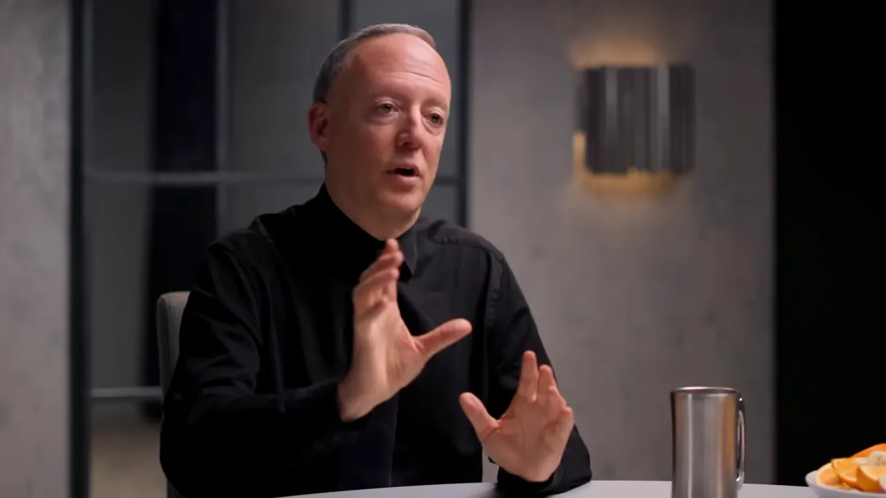 Guest seated at a table making explanatory hand gestures, stainless mug and plate of fruit visible on the table.