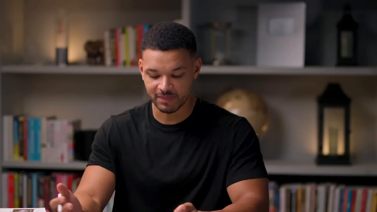 Clear mid-shot of the host seated at a table, hands visible and gesturing while explaining the microbiome effects of fasting