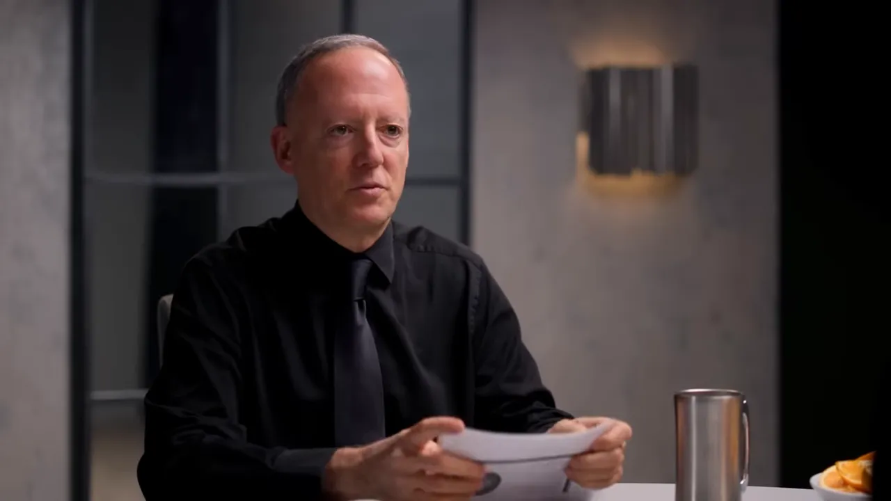 Interview guest holding a sheet of paper and speaking in a well-lit studio, head-and-shoulders view.