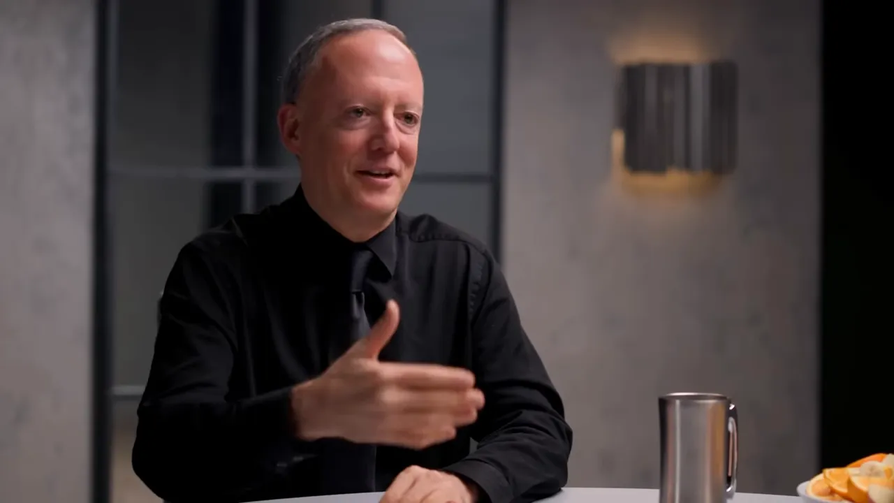 Interview guest making an open-handed gesture while describing results, with a metal mug and plate of fruit on the table