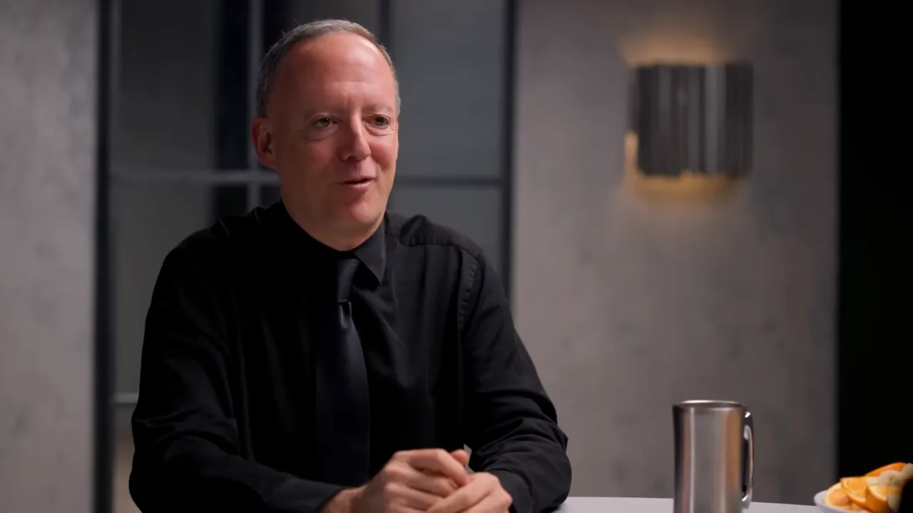 Host smiling, hands folded on the table while seated at a table with a stainless mug and a plate of sliced fruit