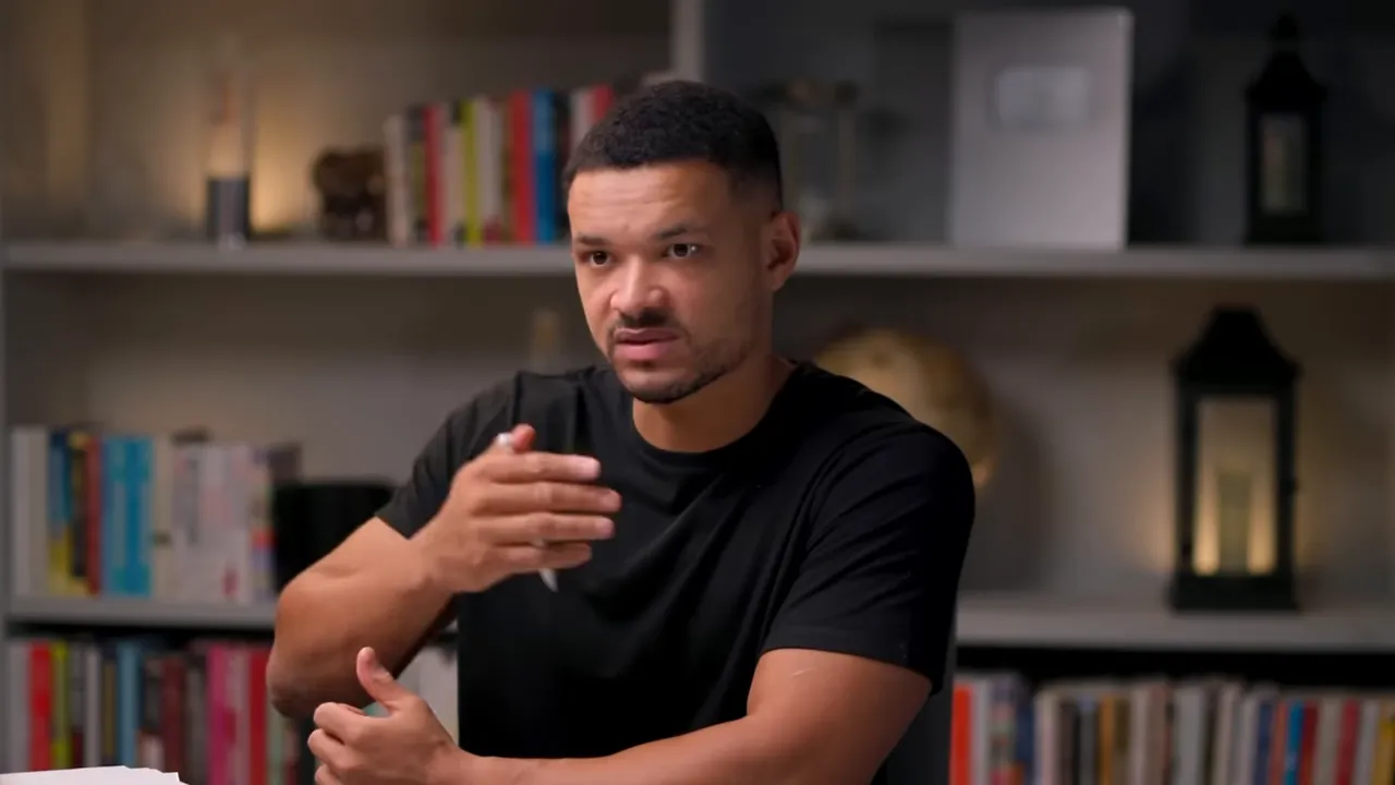 Host seated at a table gesturing while speaking in front of bookshelves, clear mid‑shot