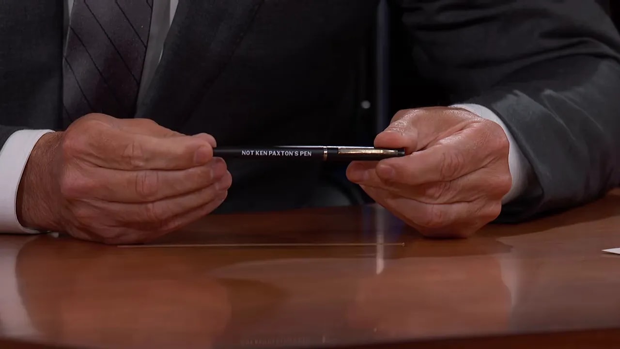 Jimmy Kimmel holding a black pen with white writing at his desk
